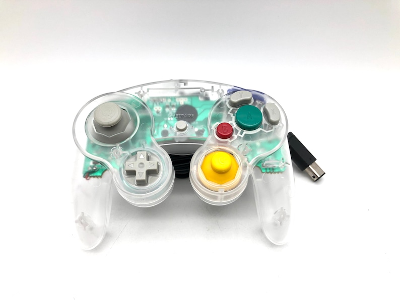 May include: A clear, translucent GameCube controller with a green and yellow button layout. The controller is connected to a black wire with a USB connector.
