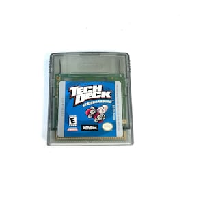 Pode incluir: Um cartucho de jogo Game Boy Color azul e branco para Tech Deck Skateboarding. O cartucho está em uma caixa de plástico transparente. O texto no cartucho diz "Tech Deck Skateboarding" e "Activision".