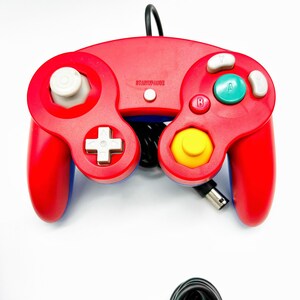 May include: A red and blue GameCube controller with a yellow button and a white start/pause button. The controller is connected to a black cord.