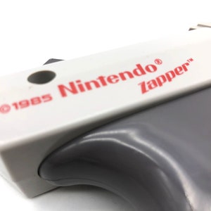 May include: A white and gray Nintendo Zapper light gun with the text "&copy;1985 Nintendo&reg; Zapper&trade;" printed on the top.