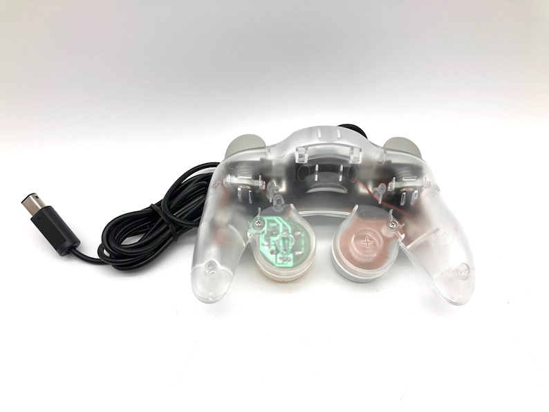 May include: A clear, transparent GameCube controller with a black cord. The controller has a green and a brown button.