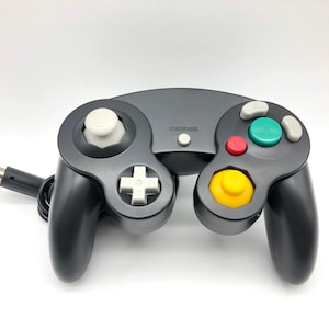 May include: A black Nintendo GameCube controller with a white control stick, a white plus button, and colored buttons. The controller has a wired connection.