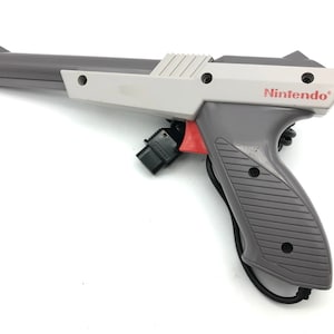 Könnte beinhalten: Eine graue und weiße Nintendo Zapper Lichtpistole für die NES-Konsole. Die Pistole hat einen roten Abzug und ein schwarzes Kabel.