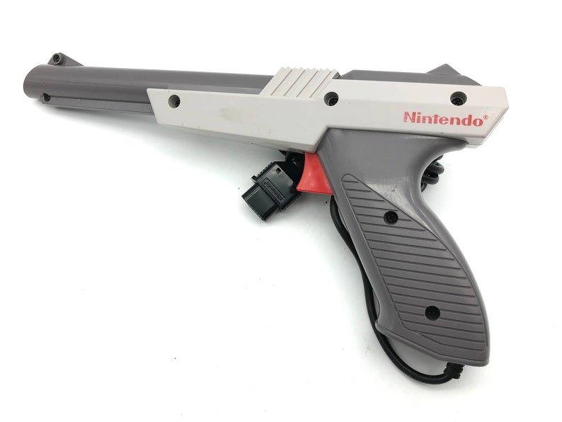 May include: A gray and white Nintendo Zapper light gun for the NES console. The gun has a red trigger and a black cord.