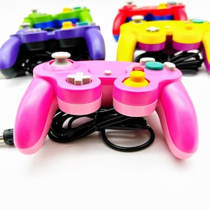 May include: A pink, purple, yellow, and green GameCube controller with a black cord. The pink controller is in focus and the other controllers are out of focus in the background.