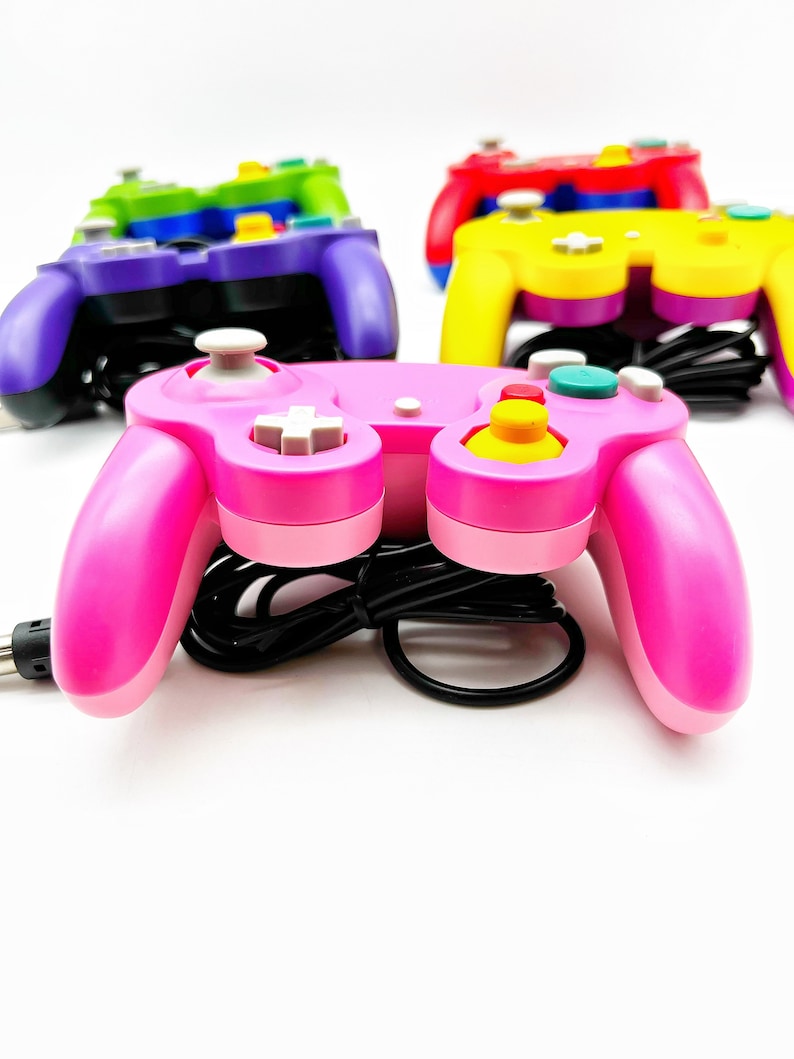 May include: A pink, purple, yellow, and green GameCube controller with a black cord. The pink controller is in focus and the other controllers are out of focus in the background.
