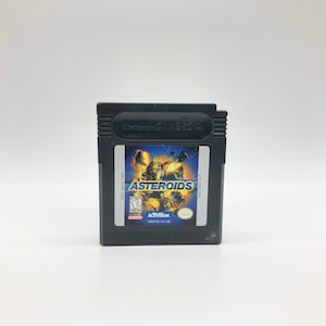 May include: A black Game Boy Color cartridge with a blue and yellow label that reads "Asteroids". The label also features a graphic of a spaceship and asteroids.