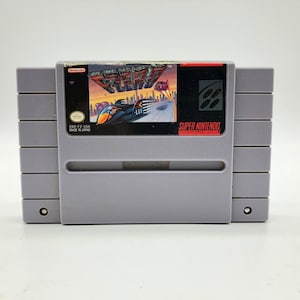 May include: A gray Super Nintendo Entertainment System game cartridge with a black label. The label features a colorful image of a futuristic race car and the title "F-Zero" in red letters. The game is for the Super Nintendo Entertainment System.
