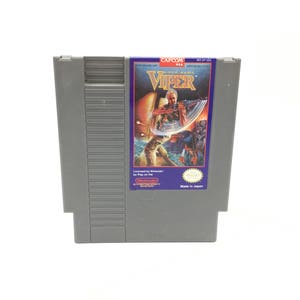 以下が含まれることがあります： カプコン製のゲーム『ザ・マーク・オブ・ザ・バイパー』のグレーの任天堂エンターテイメントシステム（NES）ゲームカートリッジ。カートリッジには、銃を持った男性の画像が描かれた紫と青のラベルが付いています。