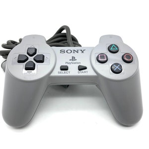 May include: A gray Sony PlayStation controller with a black directional pad, four black face buttons, and two gray buttons labeled "Select" and "Start".