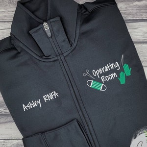 May include: Black zippered jacket with green embroidery that says "Operating Room" with a pair of scissors, a face mask, and a pair of surgical gloves. The text "Ashley RNFA" is embroidered on the left side of the jacket.