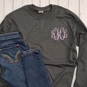 May include: A charcoal gray long-sleeve crew neck shirt featuring a light pink embroidered monogram with the letters EHL on the left chest, paired with dark blue denim jeans that have yellow stitching on the back pockets.