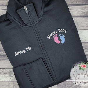 May include: Black zippered jacket with white embroidery that reads "Mother Baby" with pink and blue footprints. The jacket also has white embroidery that reads "Ashley, RN".
