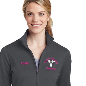 May include: A charcoal grey women's zip-up jacket with a pink embroidered logo that says "Speech Language Pathology" and a caduceus symbol. The name "Krista" is embroidered on the left side of the jacket.