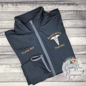 May include: A dark gray zippered jacket with the text "Respiratory Therapy" and a caduceus symbol embroidered on the right side. The text "Crystal, RRT" is embroidered on the left side of the jacket.