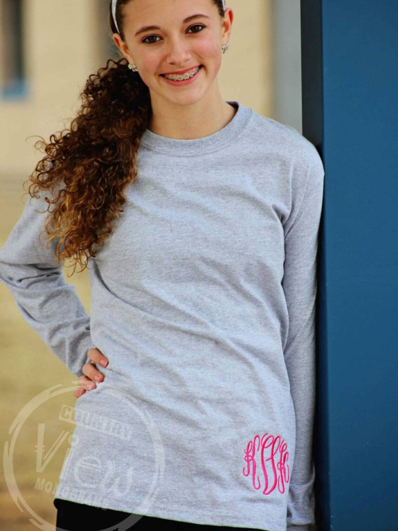 May include: A gray long-sleeved t-shirt with a pink monogram on the right side. The shirt is made of a soft, comfortable material.