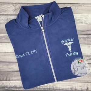 May include: A navy blue fleece zip-up jacket featuring a white embroidered caduceus symbol above the text Physical Therapy and the name Stacie, PT, DPT embroidered on the opposite side. The jacket has a high collar and a silver zipper.