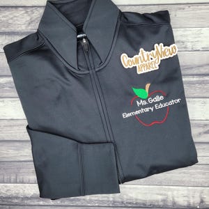 May include: A black zip-up jacket featuring a high collar and long sleeves. Embroidered on the chest is a red apple outline with a green leaf and the text "Ms. Galle Elementary Educator". A "CountryView APPAREL" logo is printed above the apple.