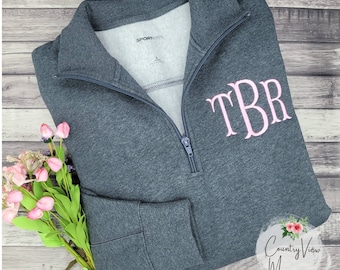 May include: A dark gray 1/4 zip sweatshirt with a pink monogram "TBR" embroidered on the front. The sweatshirt has a zippered collar and a pocket on the left sleeve. The sweatshirt is photographed on a woodgrain background.