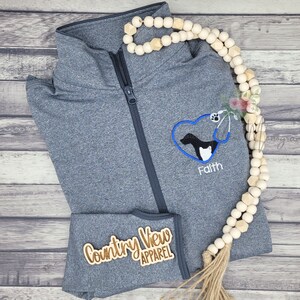May include: A grey zipped sweatshirt with a heart-shaped embroidered design featuring a dog and a stethoscope. The text "Faith" is embroidered below the heart. The sleeve has a brown wooden patch with the text "Country View Apparel".