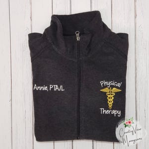 May include: A charcoal grey zip-up jacket featuring a high collar and a full-length front zip. The left side is embroidered with the text Annie, PTA/L in white, while the right side displays a gold caduceus symbol with Physical Therapy text.
