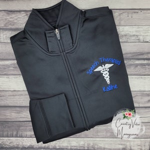 May include: Black zip-up jacket with a blue embroidered design that says "Speech Therapist" with a caduceus symbol and the name "Kathie" underneath. The jacket is folded on a woodgrain surface.