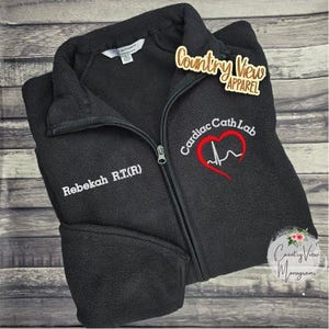 Peut inclure: Veste polaire noire zippée avec le texte "Rebekah R.T.(R)" sur la poitrine gauche. La poitrine droite présente "Cardiac Cath Lab" avec un cœur rouge et un motif ECG. La veste porte également le logo "Country View Apparel".