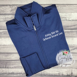 May include: A navy blue zippered jacket with white embroidery that reads "Ashley BSN RN Optional Second Line" and a white logo that reads "Country Vidor Monograms".