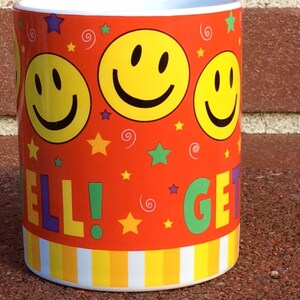 May include: A ceramic mug with a vibrant orange background, adorned with yellow smiley faces, stars, and the words "WELL! GET!". The mug has a white interior and a striped pattern at the base.