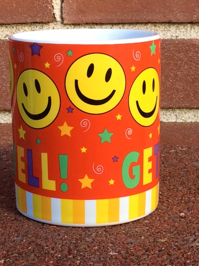 May include: A ceramic mug with a vibrant orange background, adorned with yellow smiley faces, stars, and the words "WELL! GET!". The mug has a white interior and a striped pattern at the base.