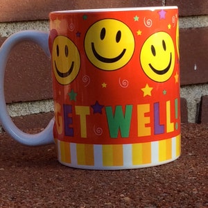May include: A ceramic mug with a white handle and rim, featuring a vibrant orange background. The mug is decorated with yellow smiley faces, stars, and the words "GET WELL!" in colorful letters. The mug is sitting on a brown surface.