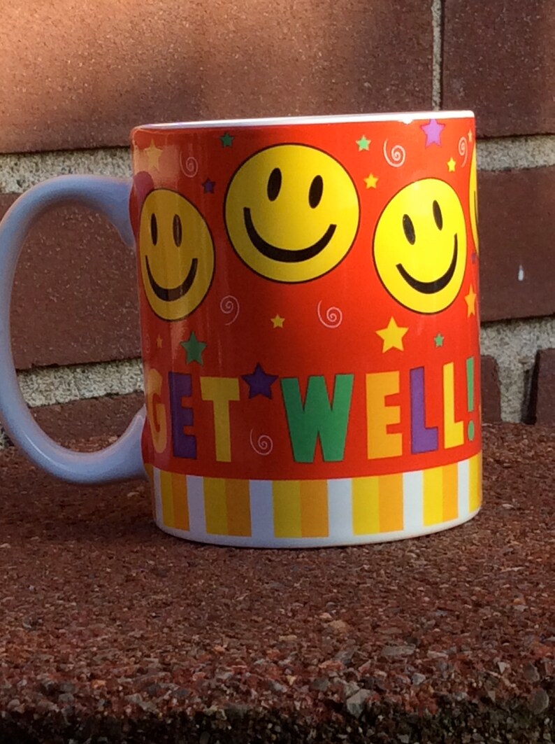 May include: A ceramic mug with a white handle and rim, featuring a vibrant orange background. The mug is decorated with yellow smiley faces, stars, and the words "GET WELL!" in colorful letters. The mug is sitting on a brown surface.