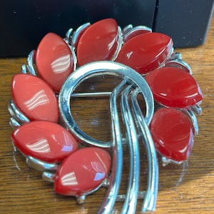 May include: A vintage brooch with a silver-tone metal frame. The brooch features eight red, oval-shaped stones arranged in a circular pattern. The design includes a central ring and stylized vertical elements.