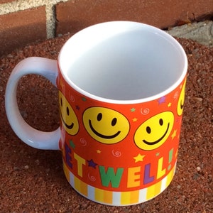 May include: A ceramic mug with a white interior and handle. The exterior is decorated with yellow smiley faces, stars, and the words "GET WELL!" against an orange background. The bottom has a striped pattern.