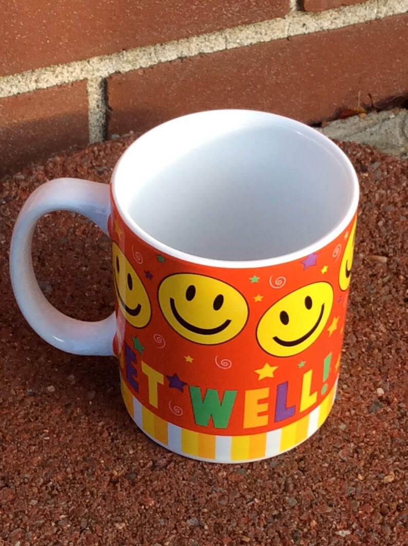 May include: A ceramic mug with a white interior and handle. The exterior is decorated with yellow smiley faces, stars, and the words "GET WELL!" against an orange background. The bottom has a striped pattern.