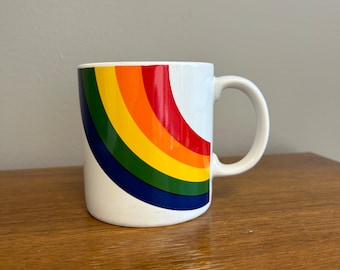 May include: White ceramic mug with a rainbow design. The rainbow is curved and has red, orange, yellow, green, blue, and indigo stripes.