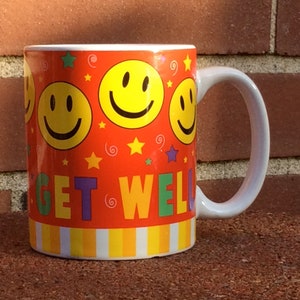 May include: A ceramic mug with a white handle and a vibrant orange background. The mug is decorated with yellow smiley faces, stars, and the words "GET WELL" in colorful letters. The bottom of the mug has a striped pattern.