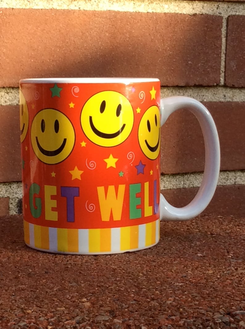May include: A ceramic mug with a white handle and a vibrant orange background. The mug is decorated with yellow smiley faces, stars, and the words "GET WELL" in colorful letters. The bottom of the mug has a striped pattern.