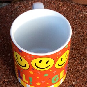 May include: A ceramic mug with a white interior and a handle. The exterior is decorated with a repeating pattern of yellow smiley faces, stars, and the words "WELL GET". The background is a red-orange color.