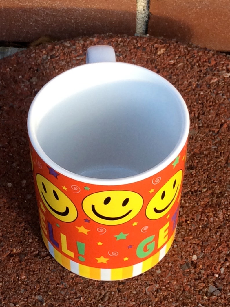 May include: A ceramic mug with a white interior and a handle. The exterior is decorated with a repeating pattern of yellow smiley faces, stars, and the words "WELL GET". The background is a red-orange color.