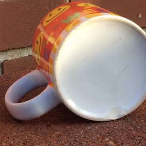 May include: Orange ceramic mug with a white interior and a light blue handle. The mug is decorated with smiling faces and colorful geometric shapes. The mug is resting on a brick surface.