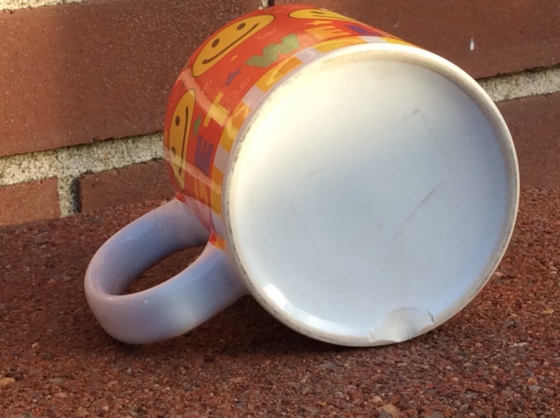 May include: Orange ceramic mug with a white interior and a light blue handle. The mug is decorated with smiling faces and colorful geometric shapes. The mug is resting on a brick surface.