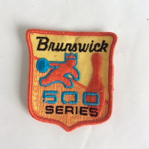 May include: A yellow and orange embroidered patch with the text "Brunswick 500 Series". The patch features a cartoon bowler throwing a bowling ball at a pin.