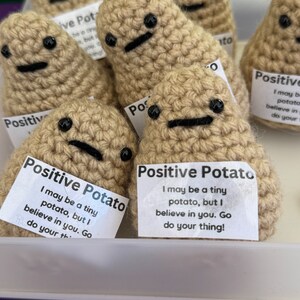 May include: A group of crocheted potato plushies with embroidered faces and the text "Positive Potato" on a white tag. The tag reads "I may be a tiny potato, but I believe in you. Go do your thing!"