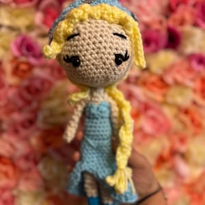 May include: A crocheted doll with blonde hair, a blue dress, and a crown. The doll is holding a long braid of yarn.