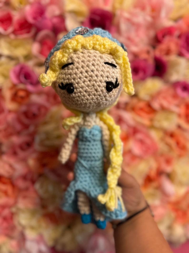 May include: A crocheted doll with blonde hair, a blue dress, and a crown. The doll is holding a long braid of yarn.