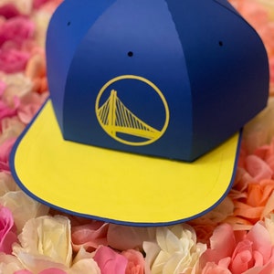 Puede incluir: Un sombrero de papel azul y amarillo con un logotipo de Golden State Warriors. El sombrero está sobre un lecho de flores rosas y blancas.