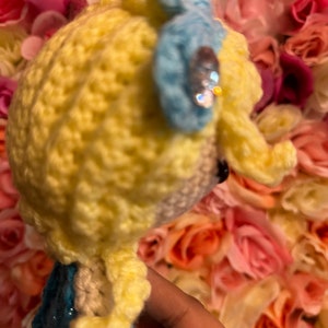 May include: A crocheted doll with yellow hair, a blue bow, and a sparkly embellishment. The doll is wearing a blue dress and is posed against a background of pink and white flowers.