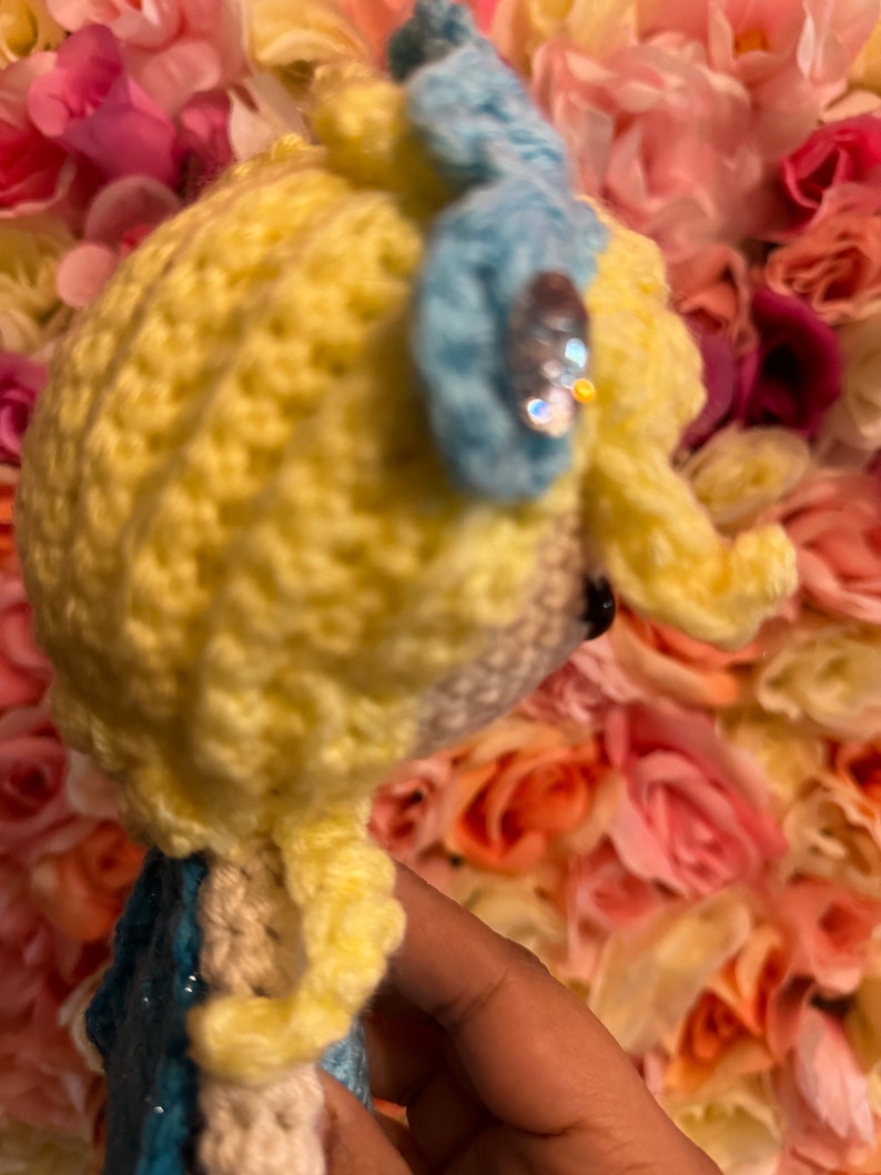 May include: A crocheted doll with yellow hair, a blue bow, and a sparkly embellishment. The doll is wearing a blue dress and is posed against a background of pink and white flowers.