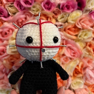 May include: A crocheted doll with a white head and black body. The head has red stitching and is covered in silver pins. The doll is standing on a hand.